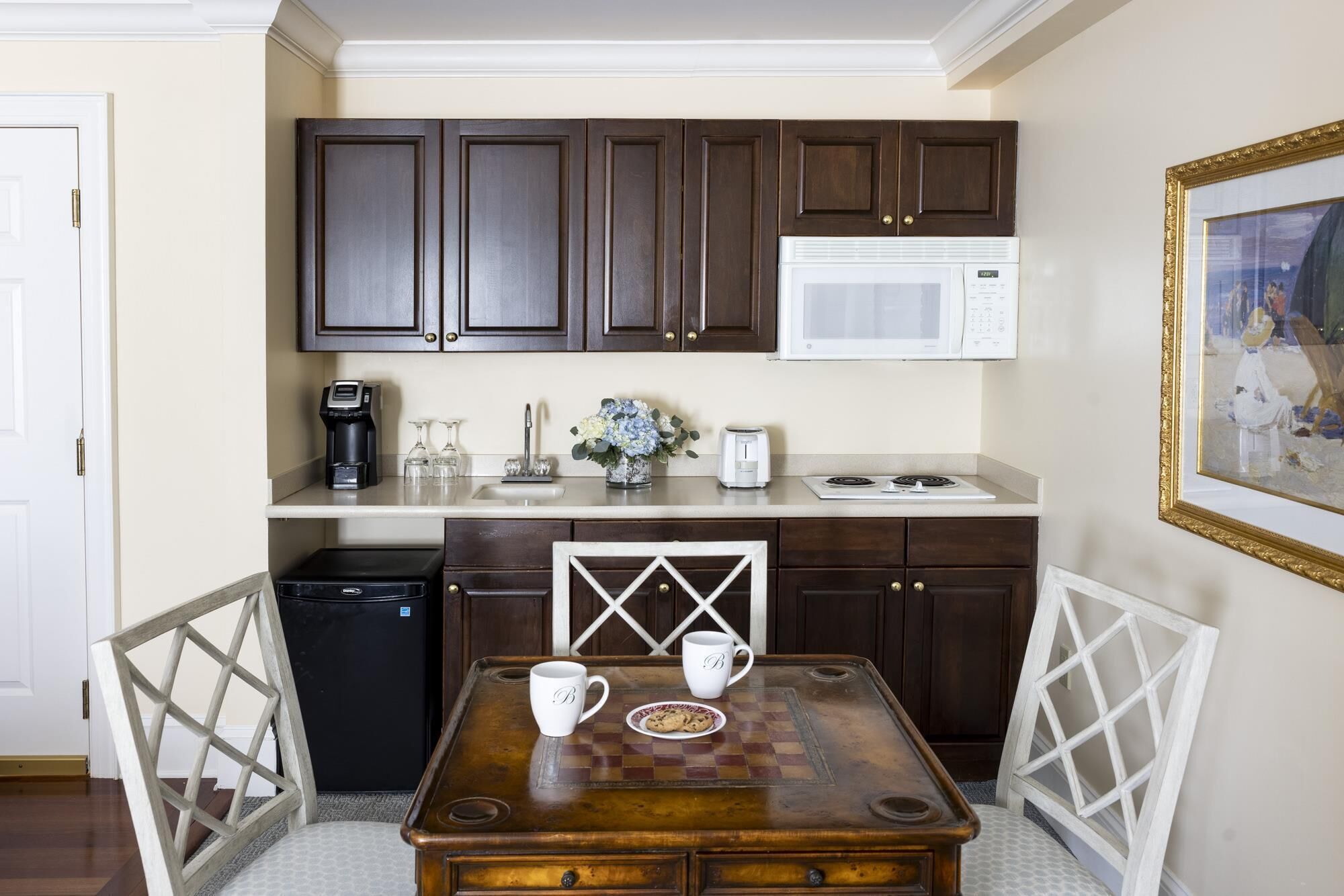 A compact kitchen with dark wooden cabinets, a white microwave, and a coffee maker. There's a small dining table with two cups and a plate of cookies.
