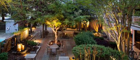 An outdoor patio area with brick flooring, wooden tables and chairs, and trees.
