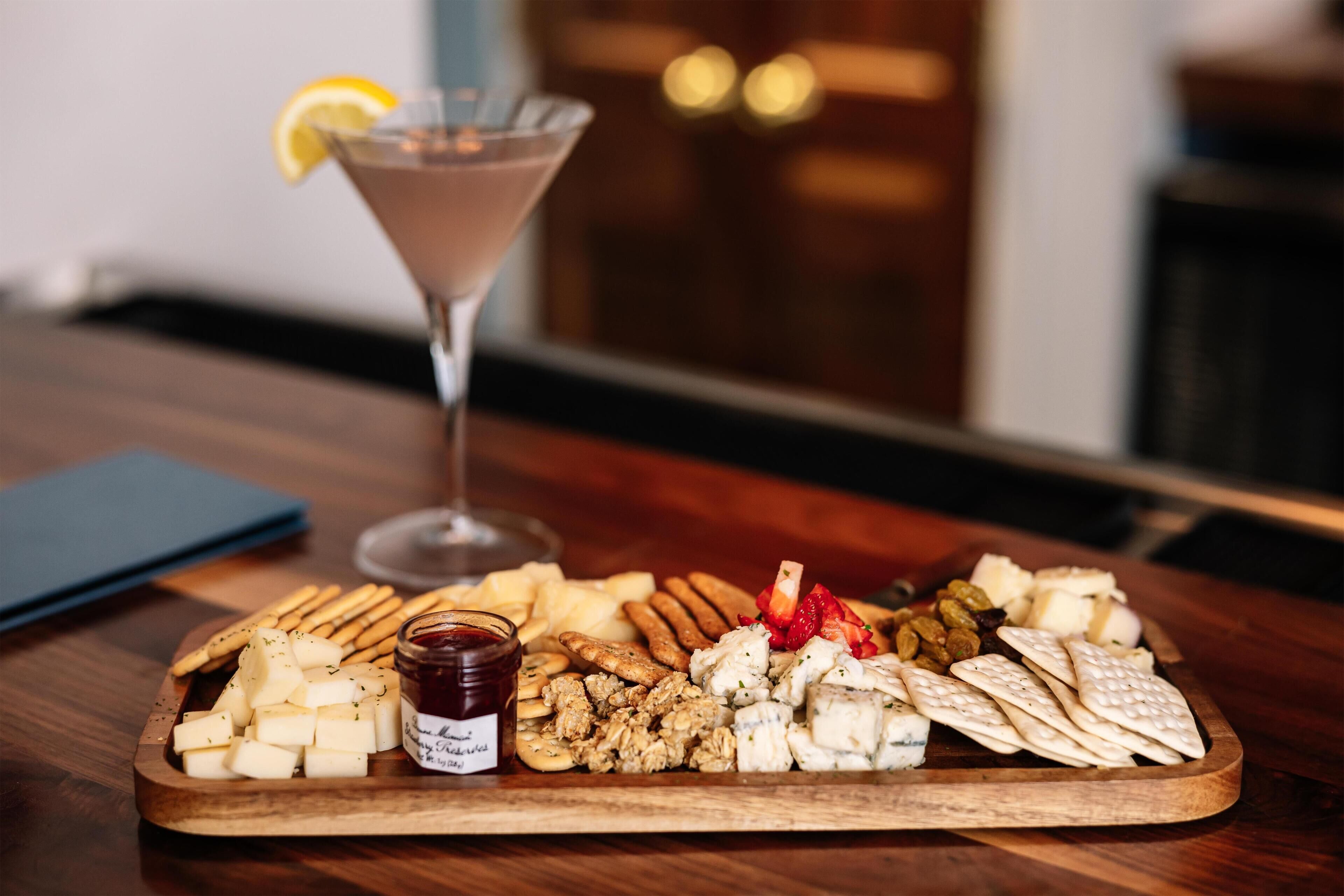 Een houten plank met diverse kazen, crackers en een pot jam. Een martini glas met een citroen is ook te zien.