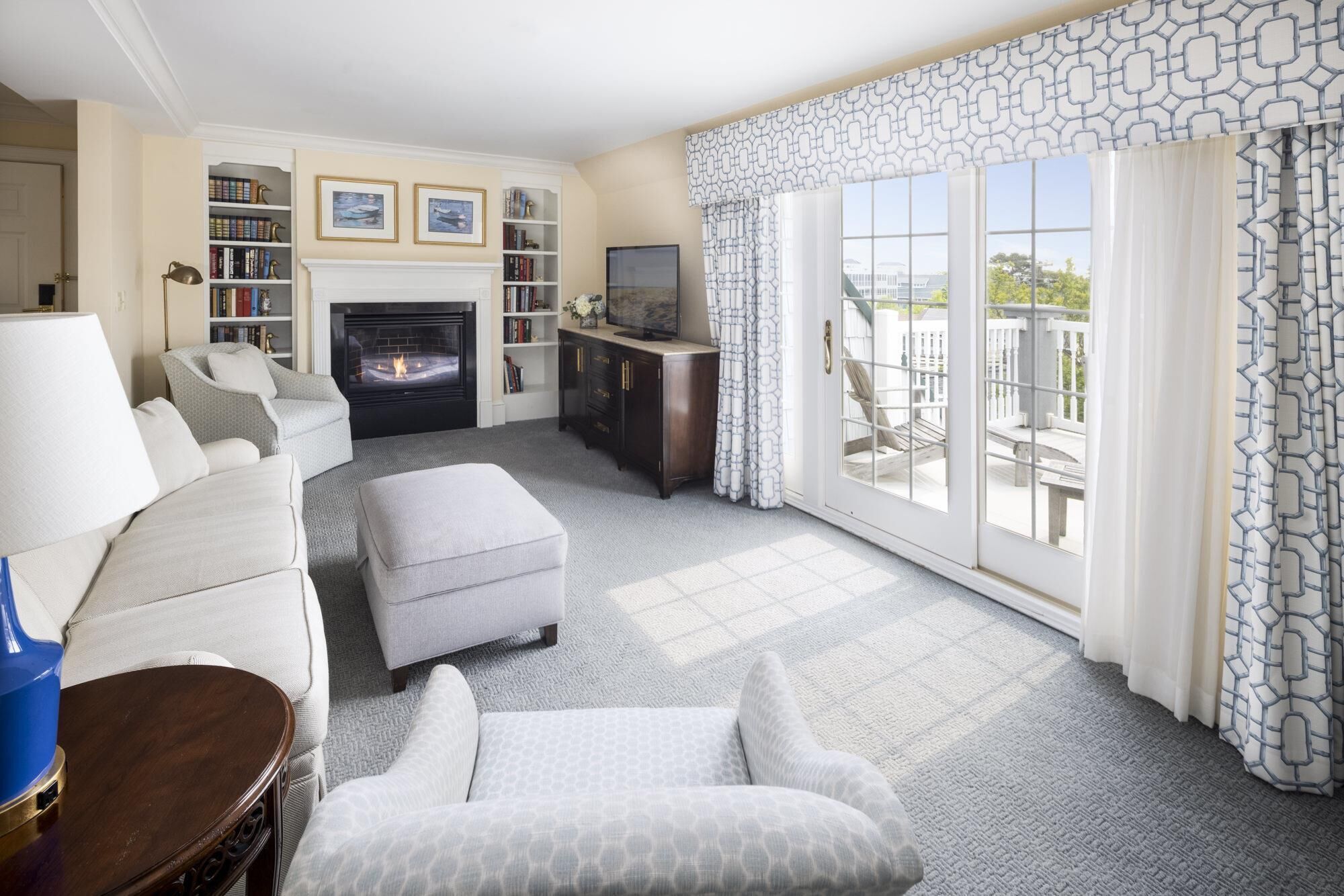 A living room with a fireplace, a sofa, an ottoman, a television, and a balcony with patterned curtains.