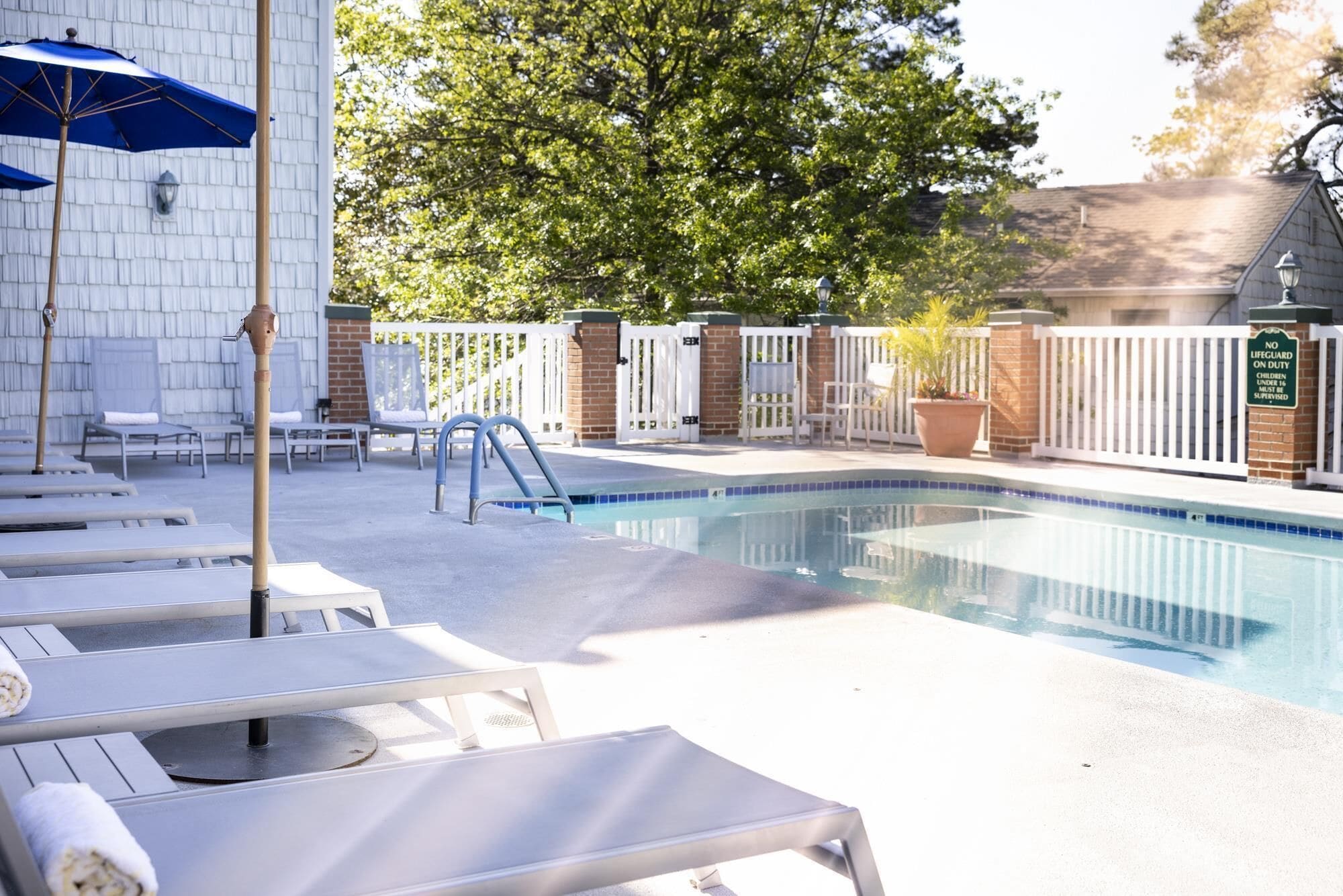 Une piscine entourée d’une clôture blanche et d’arbres, avec des chaises longues et des parasols.