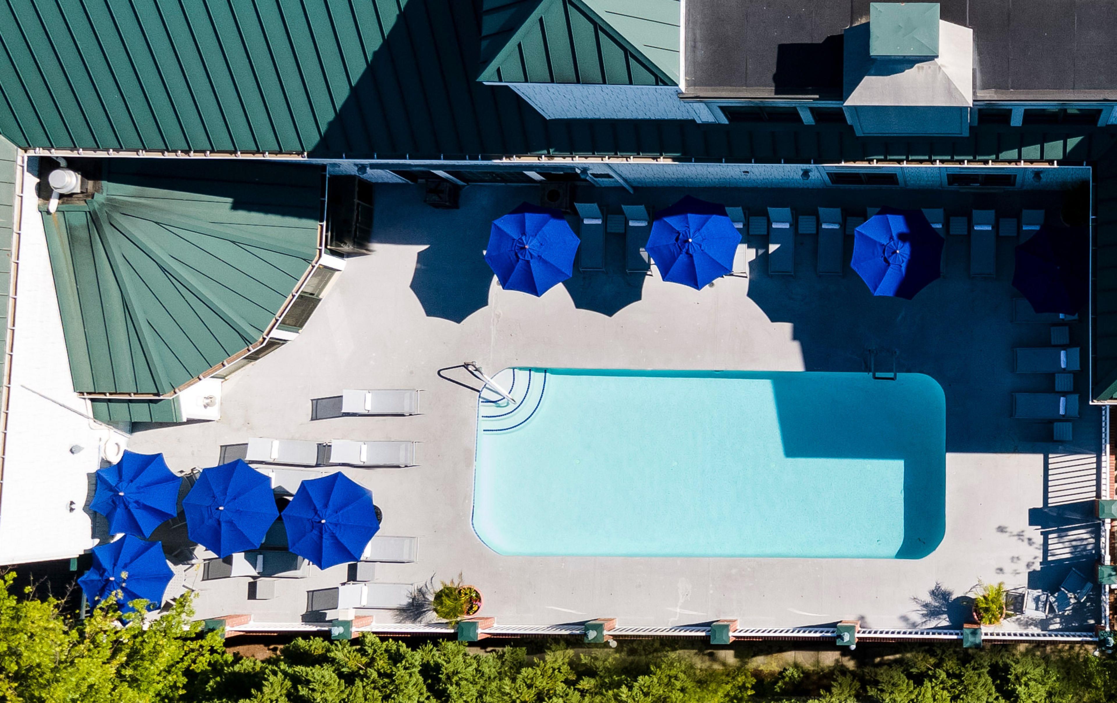 An aerial view of a hotel with a swimming pool, multiple blue umbrellas, and outdoor seating areas.