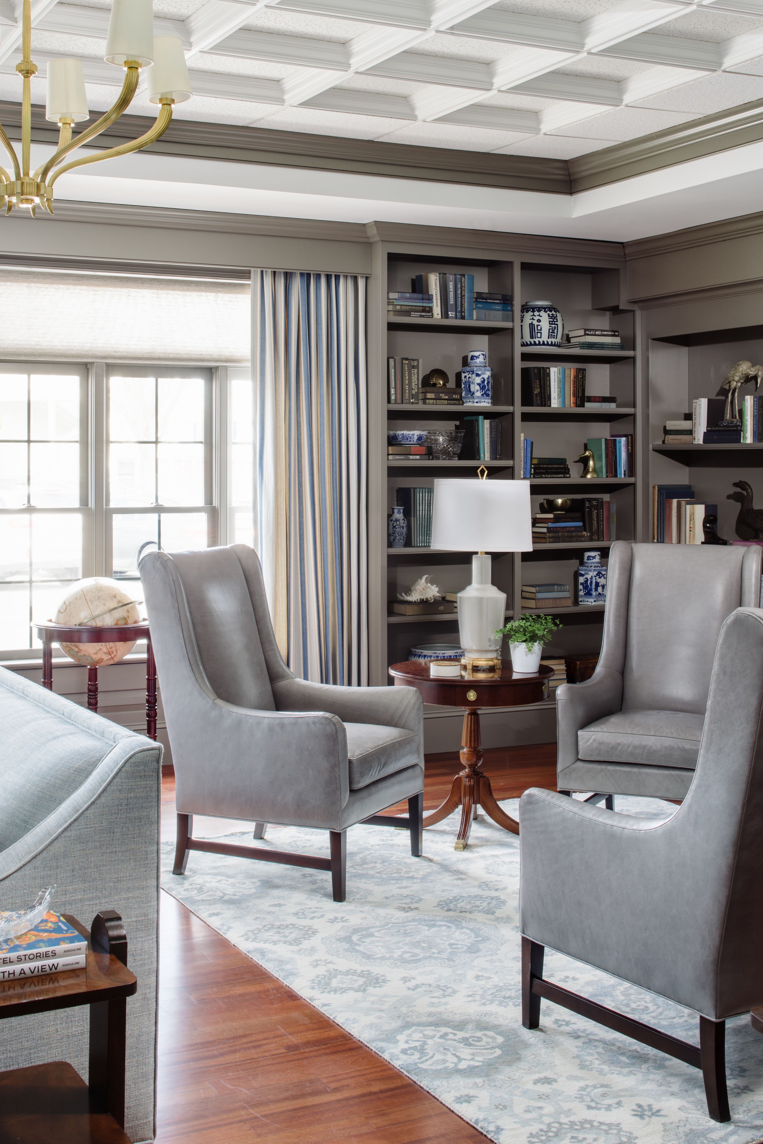 A well-lit room with a large bookshelf, two grey armchairs, a round table with a globe, and a patterned rug.