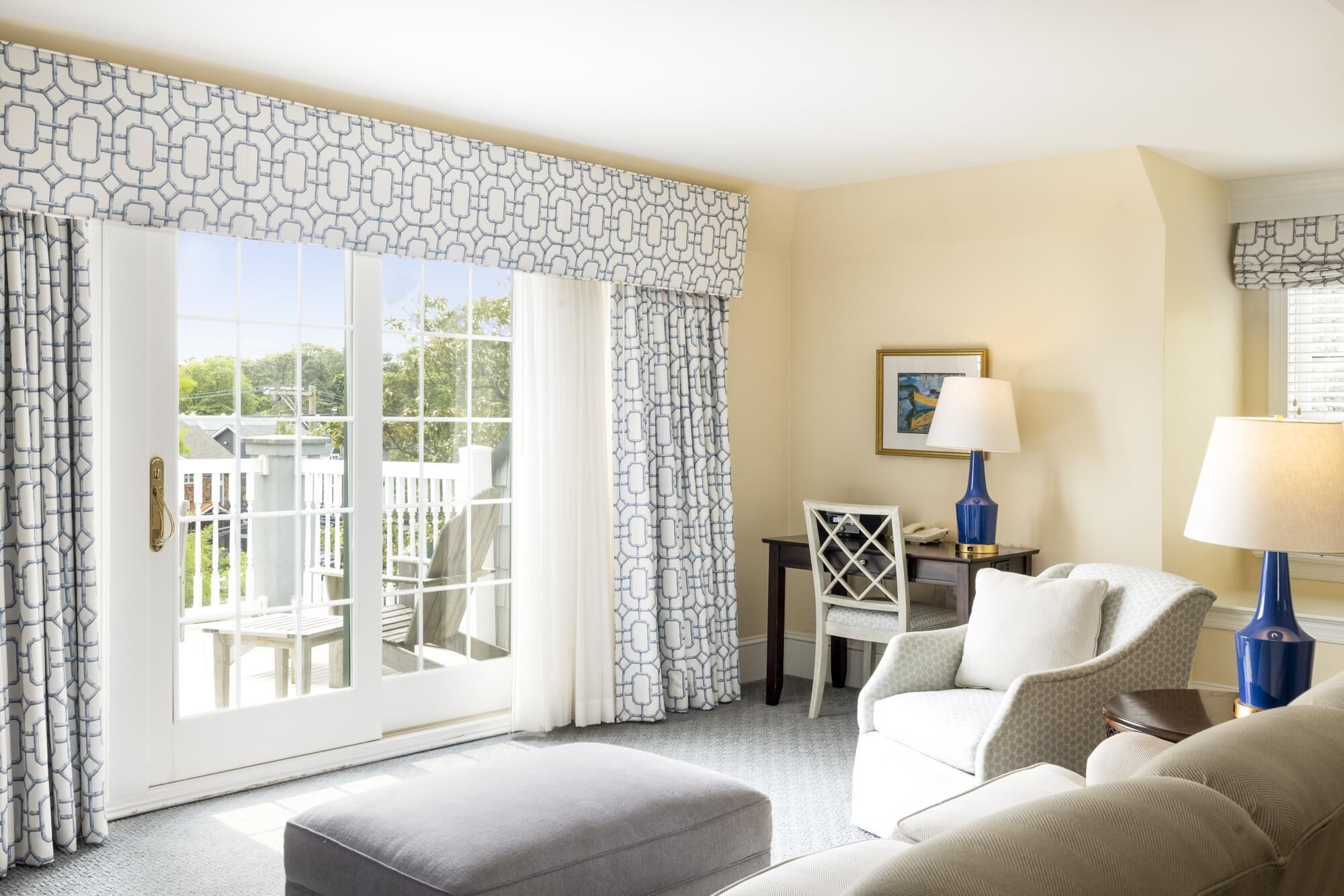 A hotel room with a large sliding glass door leading to a balcony, a desk with a chair, a sofa, and two lamps.