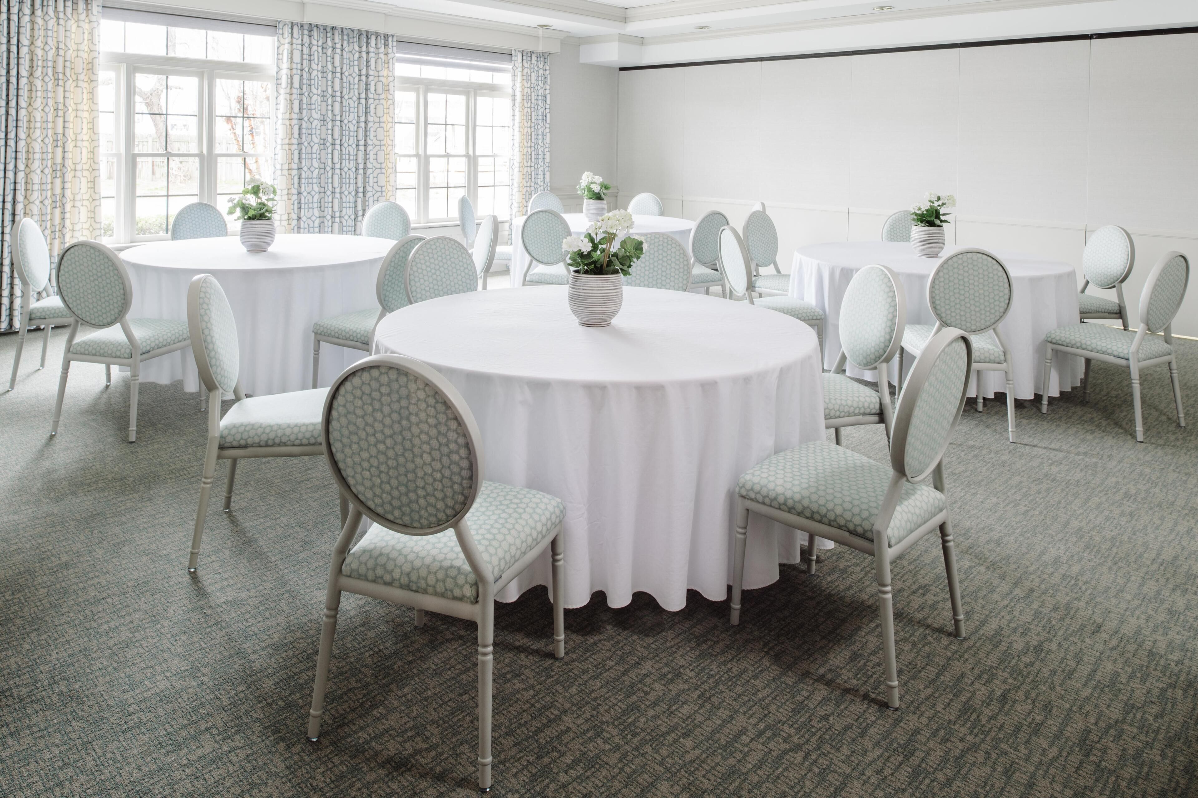 Un salón de conferencias con mesas y sillas redondas, ventanas amplias y suelo alfombrado.