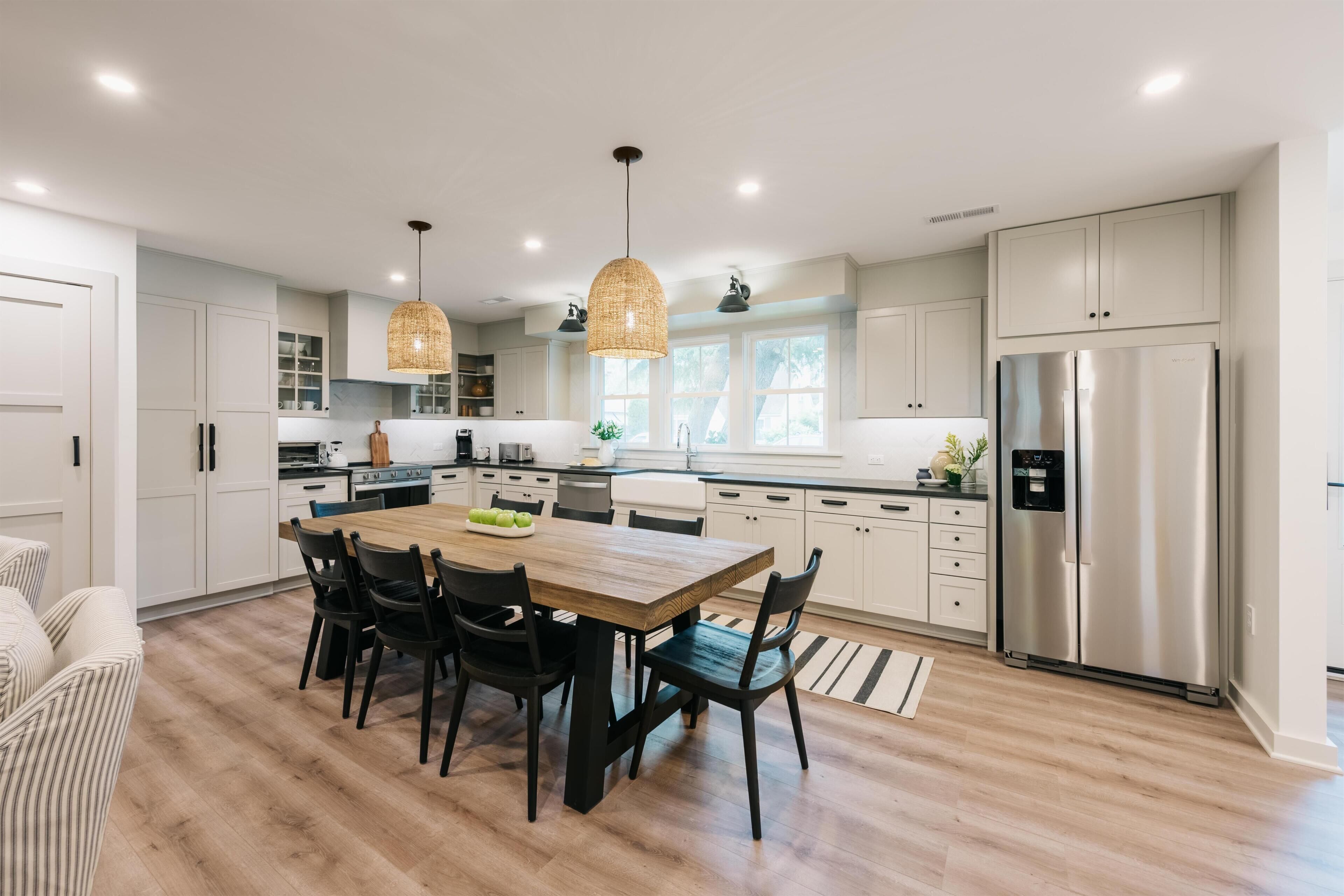 A modern kitchen with white cabinets, a stainless steel refrigerator, a wooden dining table with chairs, and pendant lighting.