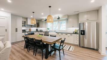 Una cocina moderna con armarios blancos, un refrigerador de acero inoxidable, una mesa de comedor de madera con sillas e iluminación colgante.