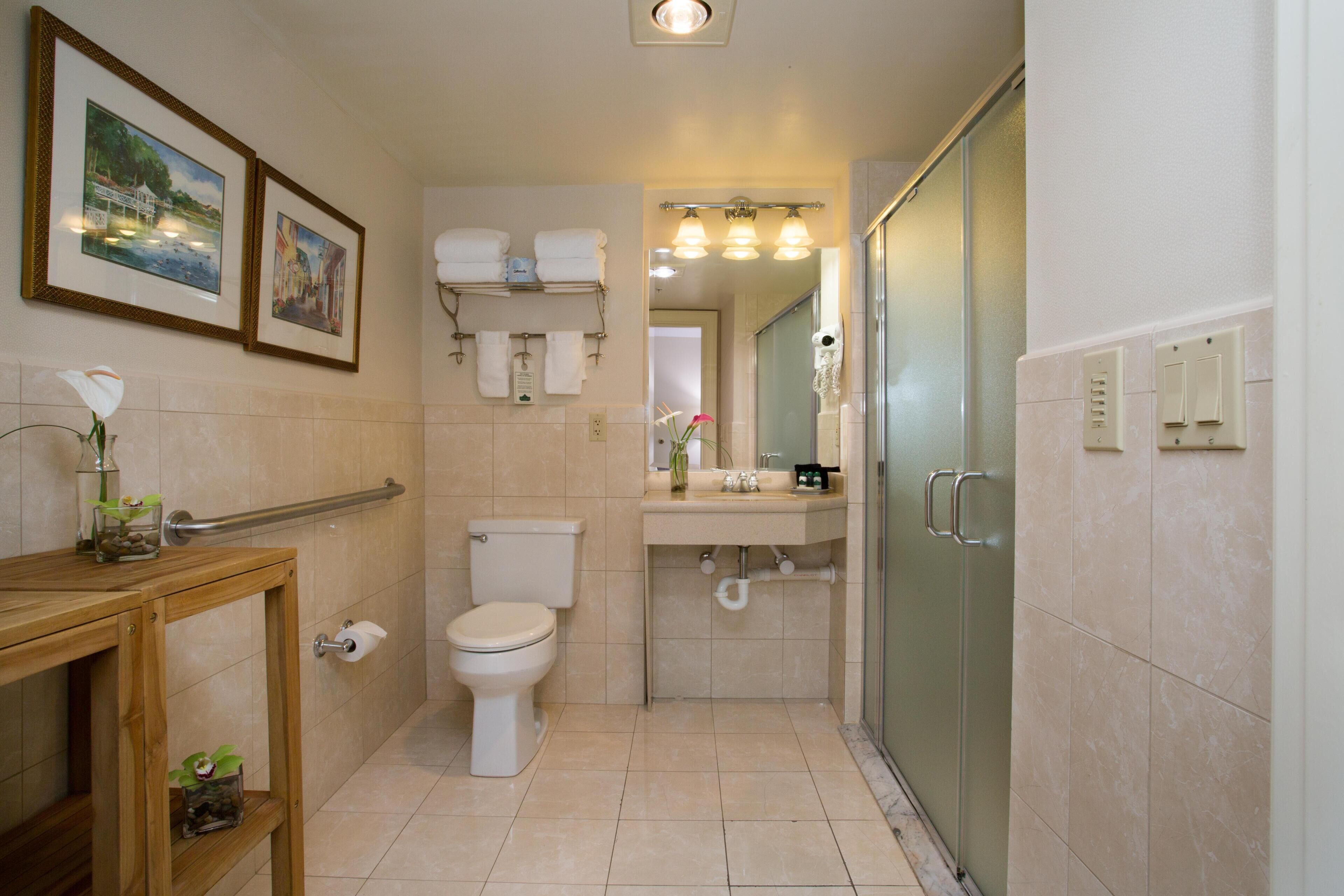 Une salle de bain avec des toilettes, un lavabo et une douche. Des tableaux encadrés ornent le mur, et une petite étagère est décorée de fleurs.