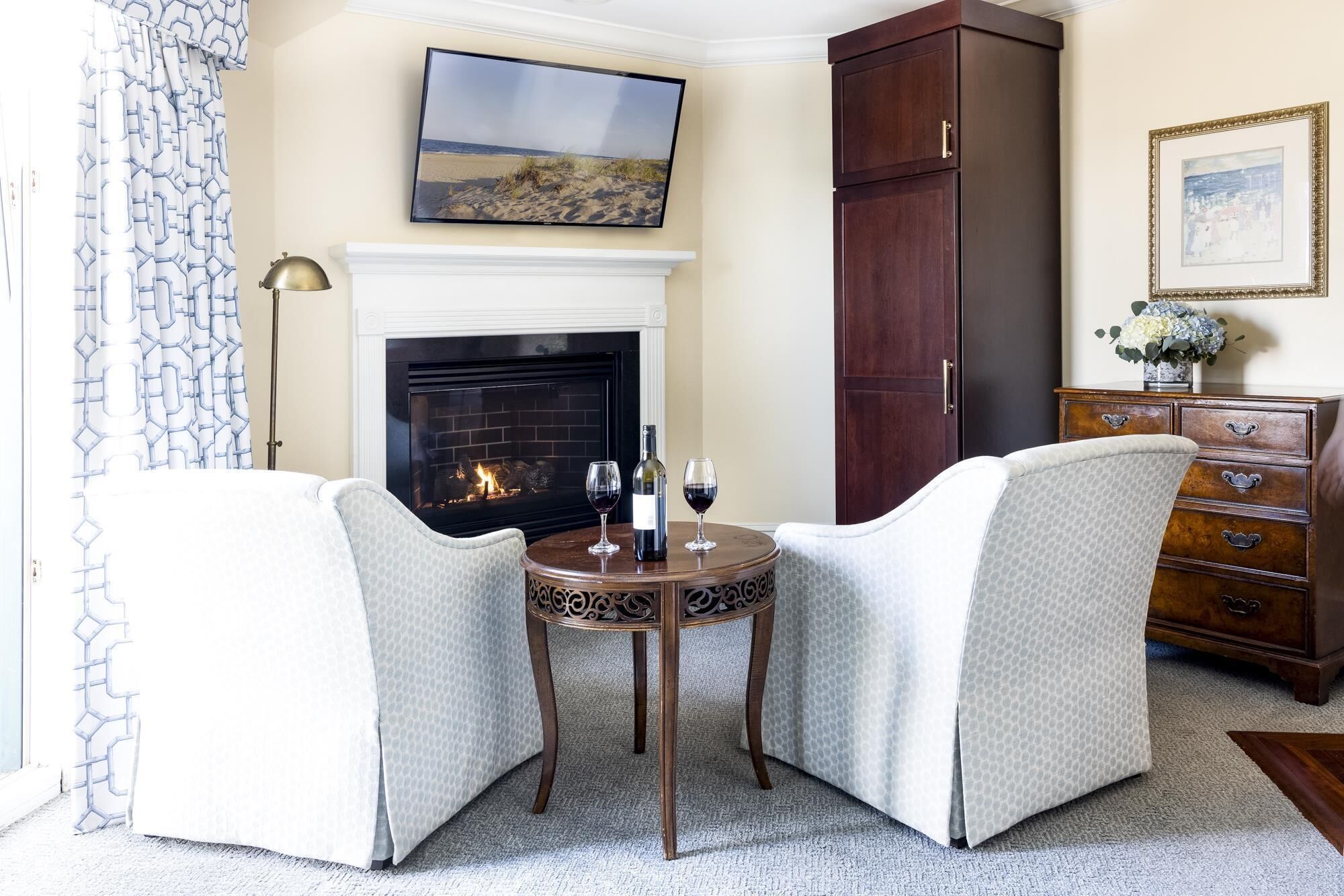 Un salón acogedor con chimenea, dos sillones, una mesita con copas de vino y una botella, y un aparador con un florero.