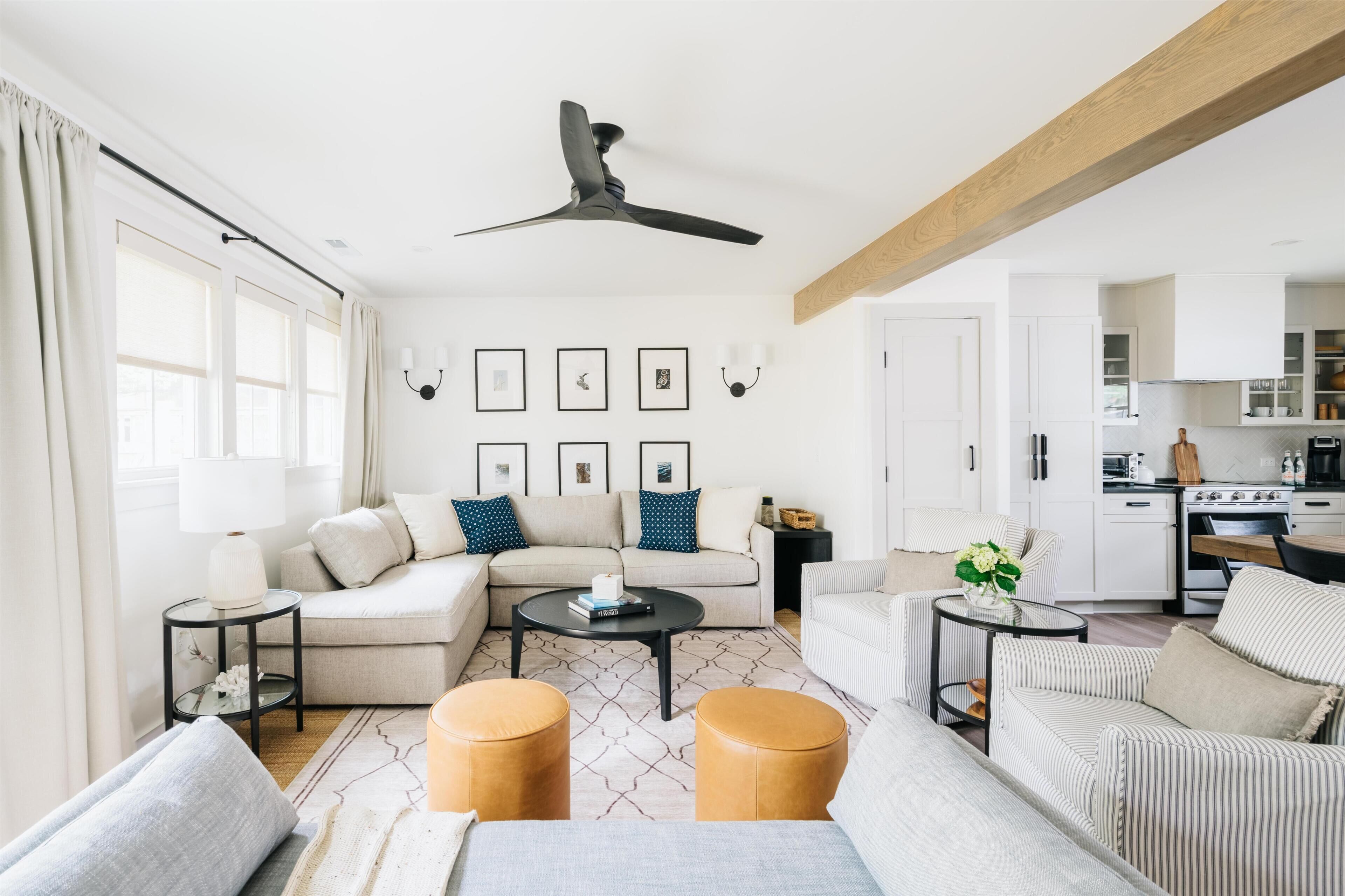 A modern living room with a sofa, coffee table, ottomans, and a ceiling fan. There is a kitchen in the background with white cabinets and a stove.