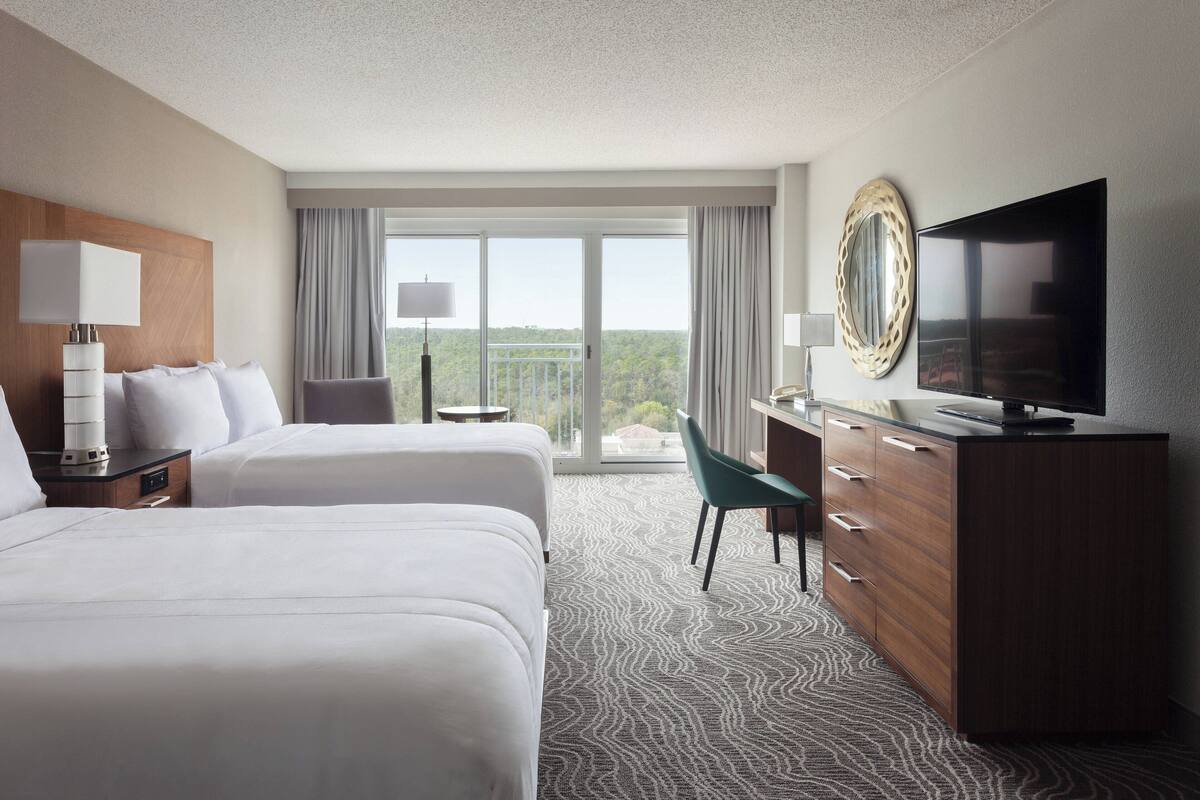 A hotel room with two beds, a TV, a desk with a chair, and a large window with a view.