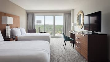 Quarto de hotel com duas camas, televisão, escrivaninha com cadeira e uma janela grande com vista.