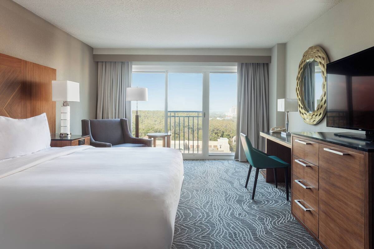 A hotel room with a large bed, a desk, a chair, a TV, and a balcony with a view.