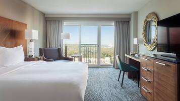 A hotel room with a large bed, a desk, a chair, a TV, and a balcony with a view.