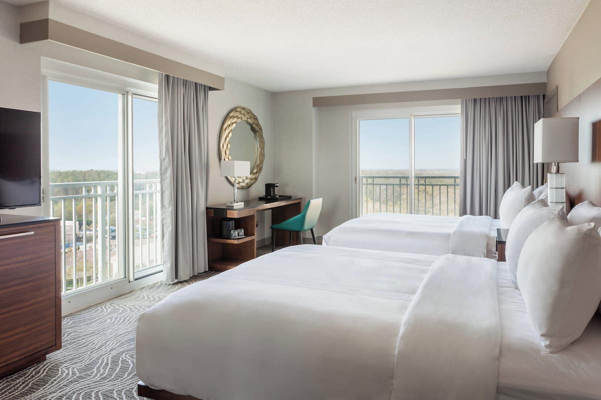 A hotel room with two beds, a desk, a chair, a TV, and large windows with curtains.