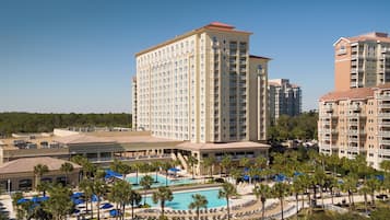 Experience the modern architecture of this high-rise hotel, featuring a central building with numerous balconies and a well-maintained pool area surrounded by palm trees.