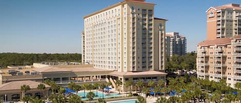 A hotel with multiple high-rise buildings, a central swimming pool surrounded by palm trees and lounge chairs, and a clear blue sky.