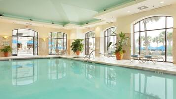 An indoor swimming pool with clear glass walls, potted plants, and lounge chairs.