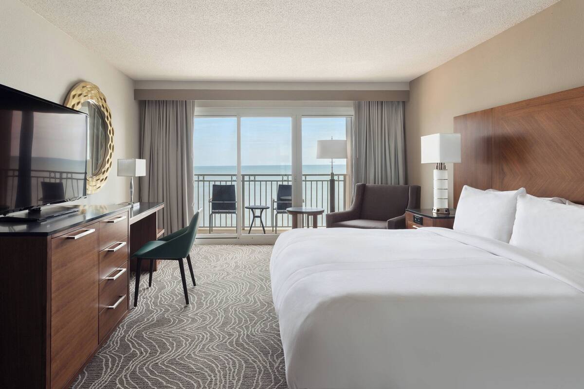 A hotel room with a large bed, a television, a desk with a chair, a small table, and a balcony with a view of the ocean.