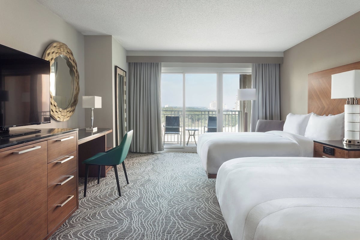 A hotel room with two beds, a desk, a chair, a TV, and a balcony with a view.