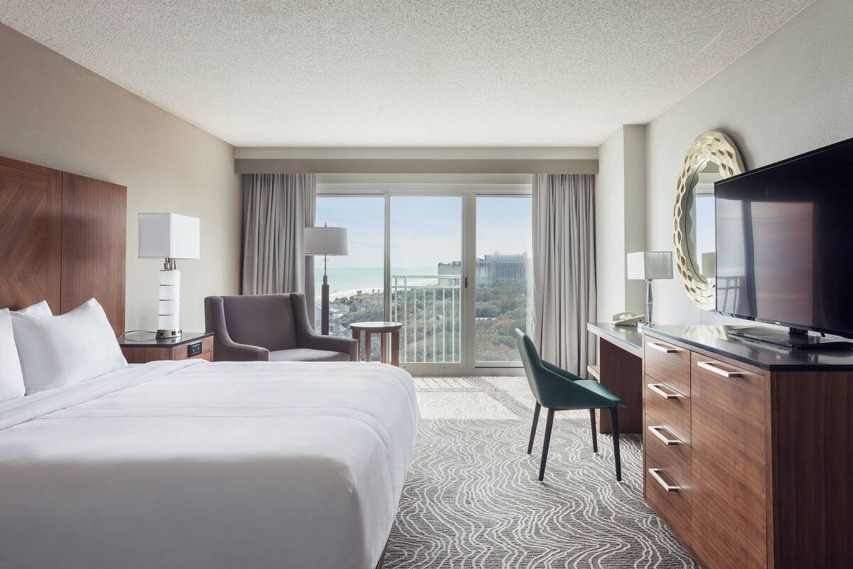 A hotel room with a large bed, a desk with a TV, a chair, a desk lamp, and a view of the city through large windows.