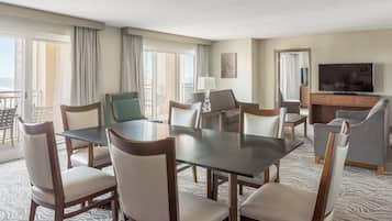 Quarto de hotel com mesa de jantar, cadeiras, sofá, televisão e varanda com cortinas.