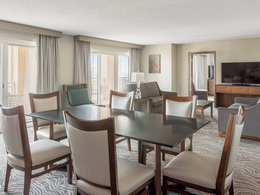 Quarto de hotel com mesa de jantar, cadeiras, sofá, televisão e varanda com cortinas.