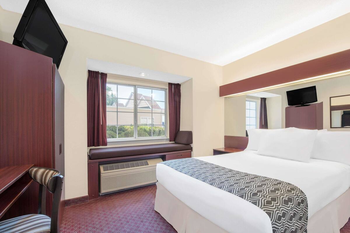 A hotel room with a bed, a desk, a chair, a window with curtains, a heater, a television, and a mirror.