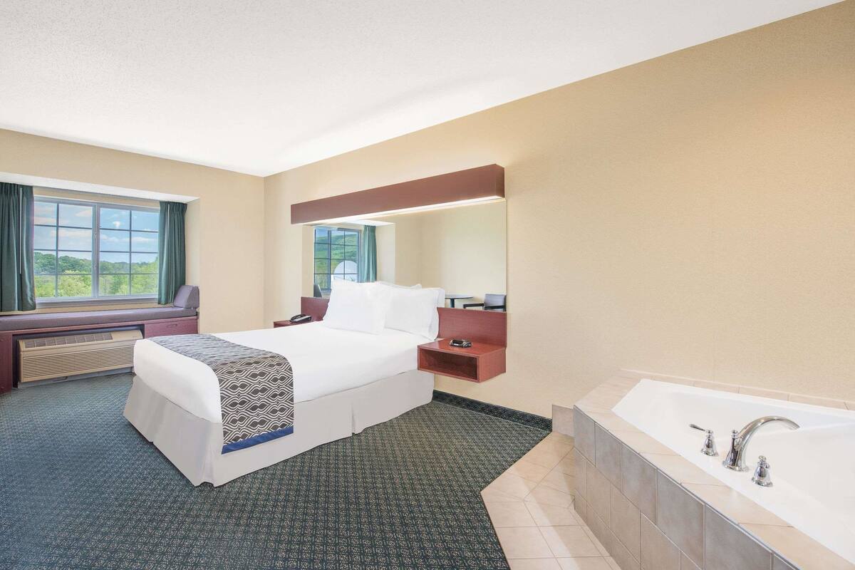 A hotel room with a bed, a desk, a chair, a large window, a bathroom with a bathtub, and a mirror.