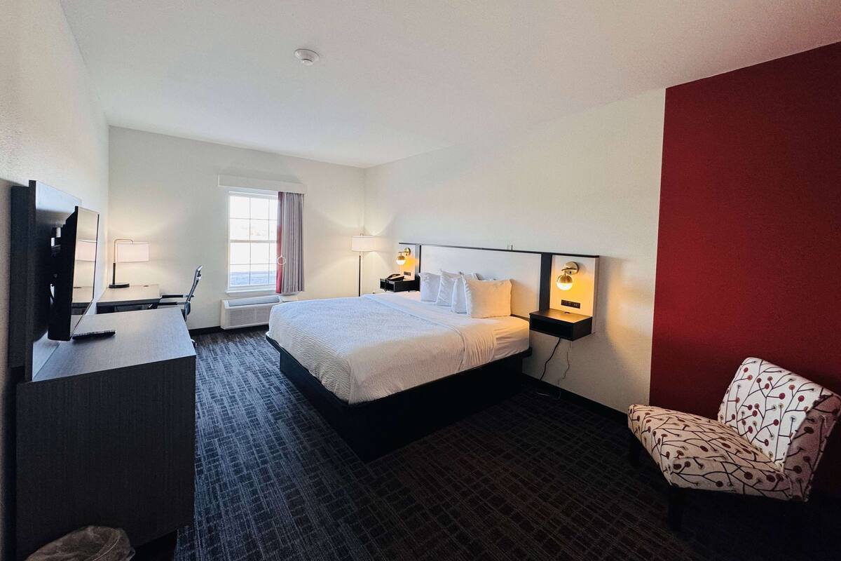 A hotel room with a bed, a desk with a TV, a chair, and a nightstand with a lamp.