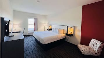 A hotel room with a bed, a desk with a TV, a chair, and a nightstand with a lamp.