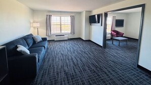 A hotel room with a dark blue sofa, a small table, a TV mounted on the wall, and a window with curtains.