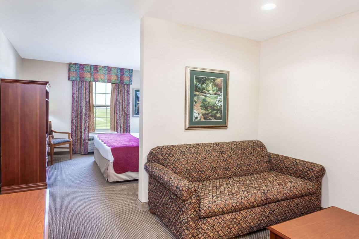 A hotel room with a patterned sofa, a bed with a red blanket, a wooden wardrobe, a framed picture on the wall, and a window with curtains.