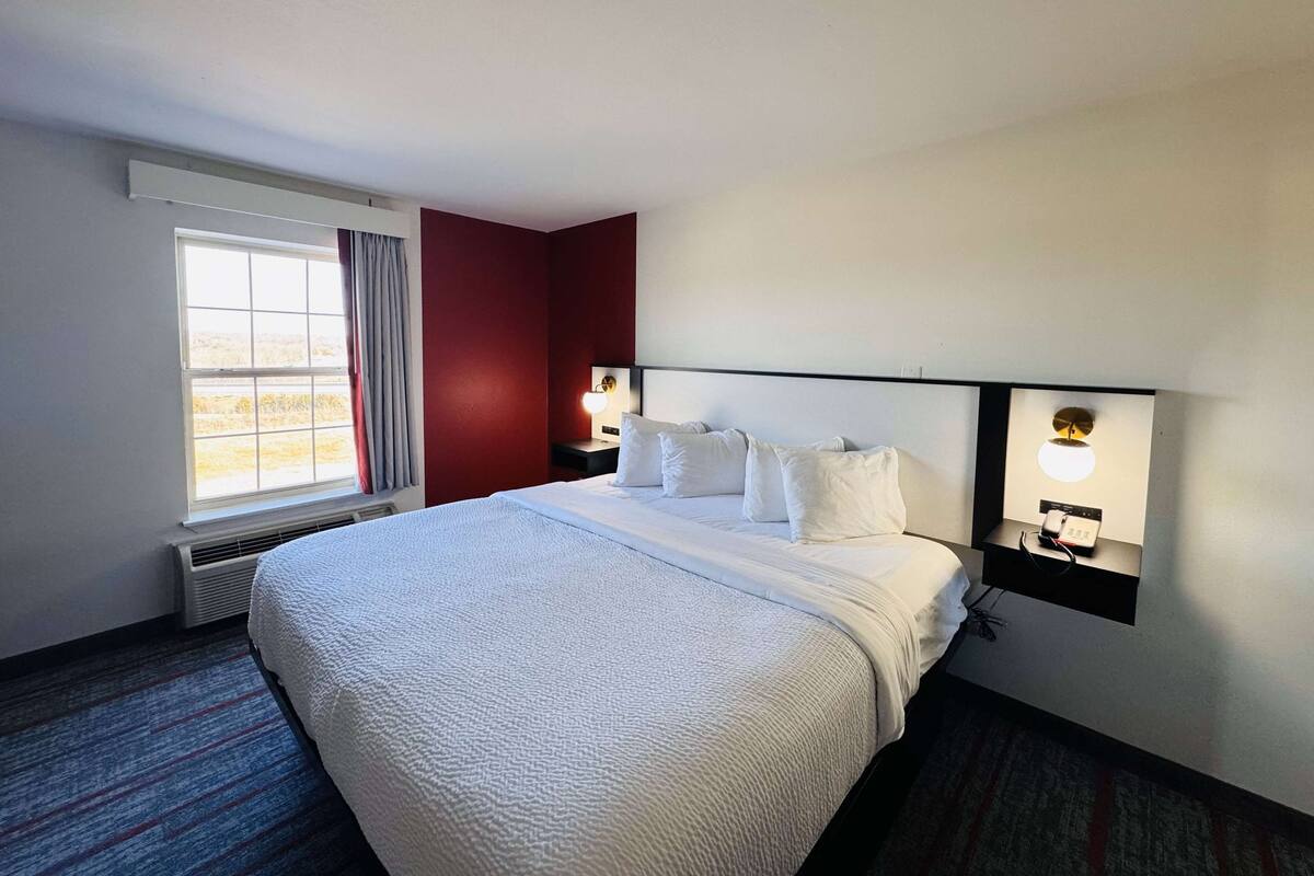 A hotel room with a large bed, two bedside tables with lamps, a window with curtains, and a red accent wall.