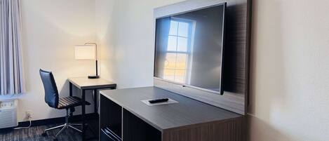 Habitación de hotel con televisión de pantalla plana montada en la pared, un escritorio con silla, una lámpara y un ventanal con cortinas.