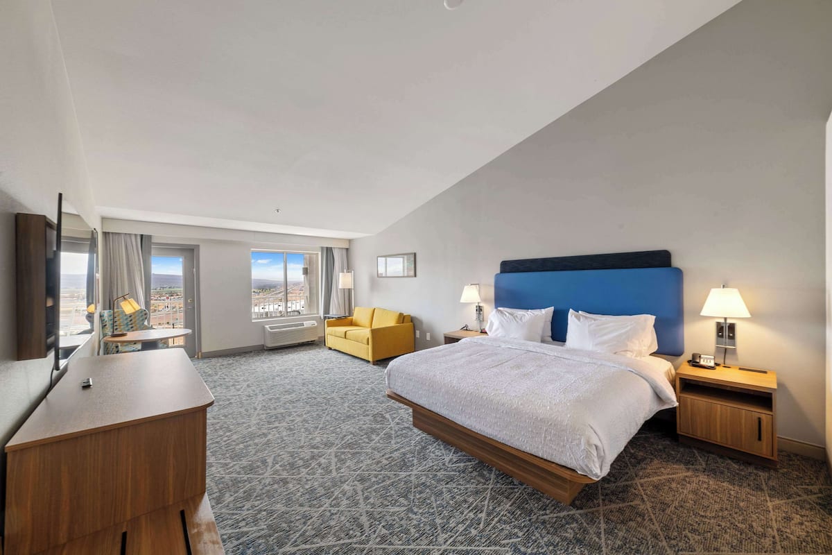 A hotel room with a large bed, a desk, a chair, a nightstand with a lamp, and a view of the city.