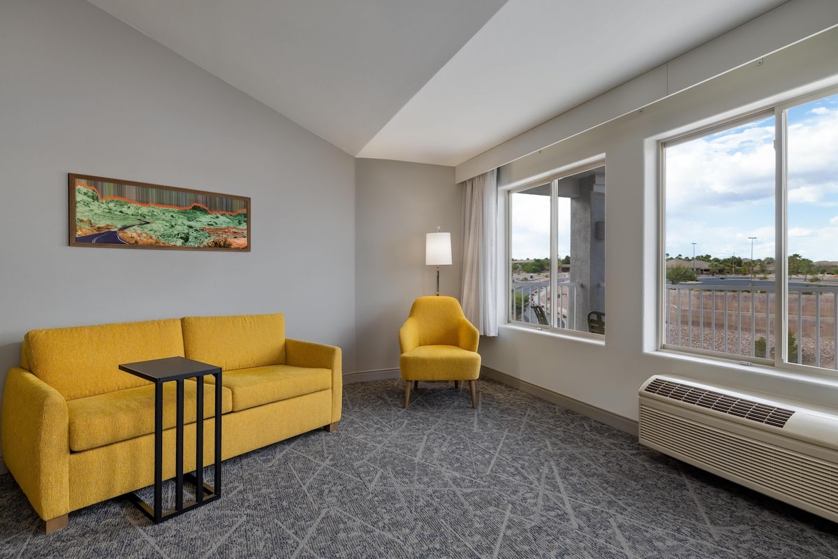 A room with a yellow sofa, a small table, a yellow chair, a painting on the wall, and a large window with a view of the outside.