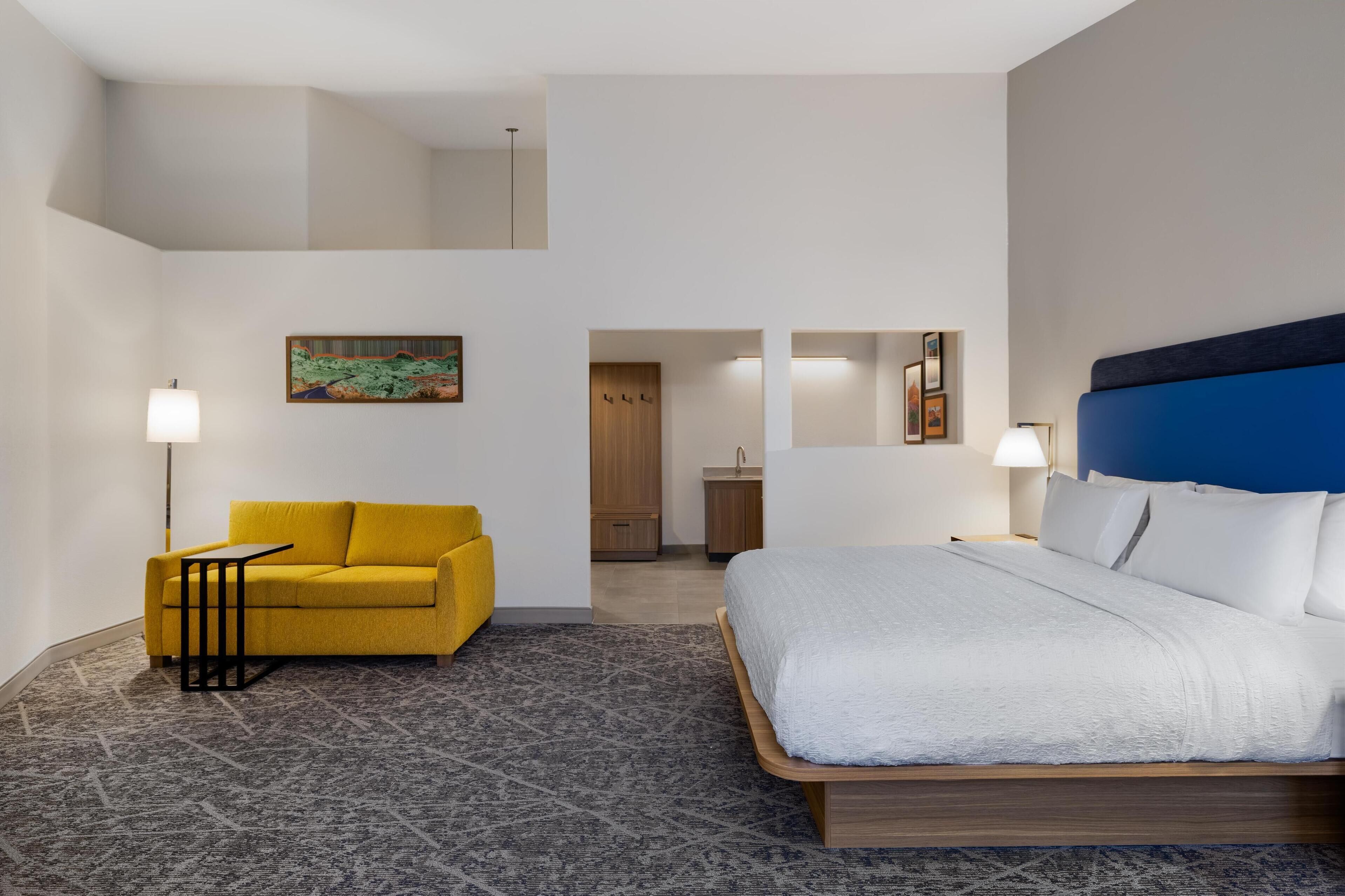Ett hotellrum med en säng, en gul soffa, ett litet bord och en tavla på väggen.