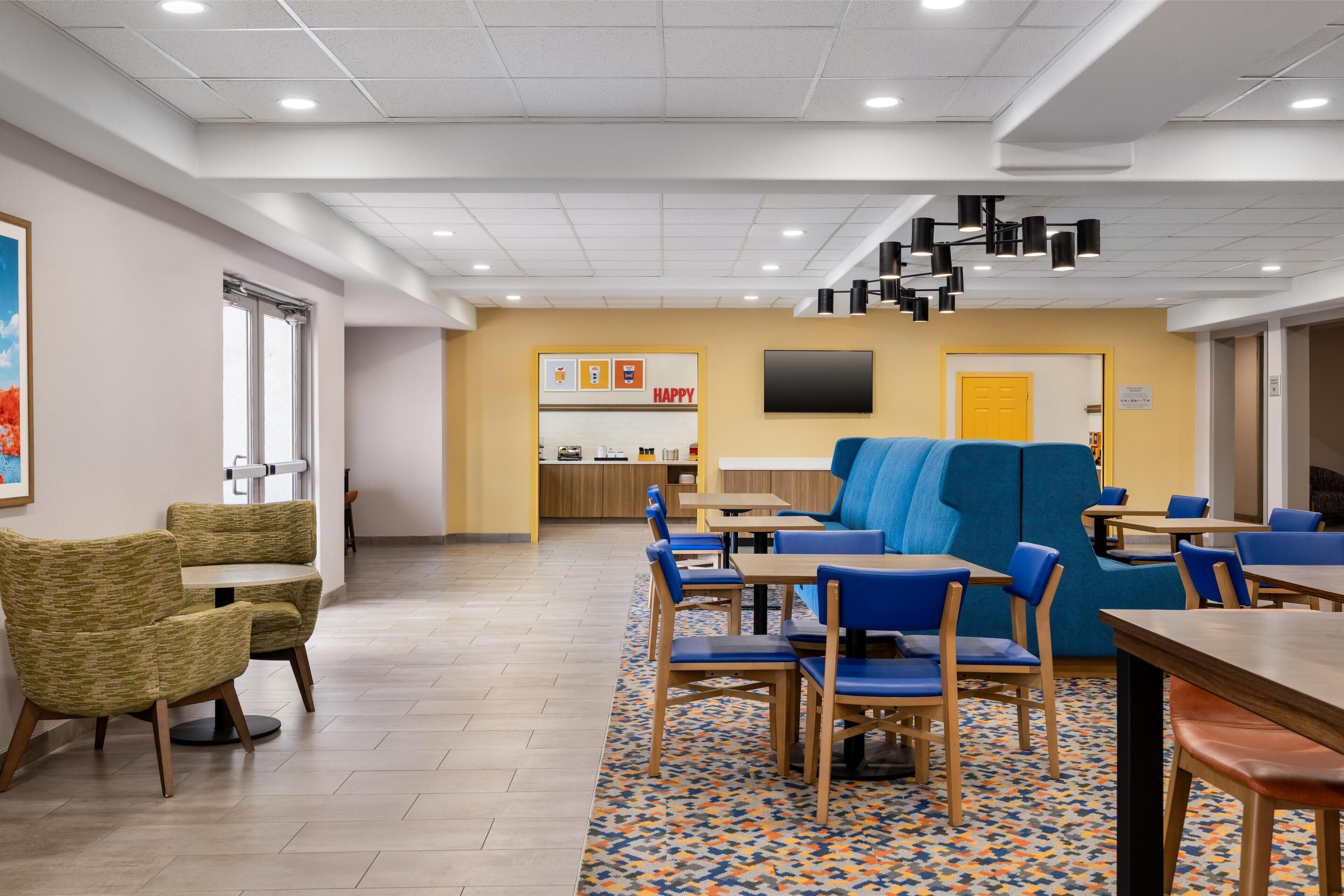 A modern dining area with multiple tables and chairs, a kitchen in the background, and a large screen on the wall.