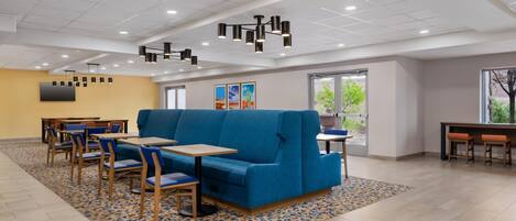 A spacious indoor area with a blue sofa, wooden tables, and chairs. There are ceiling lights and a large window providing natural light.