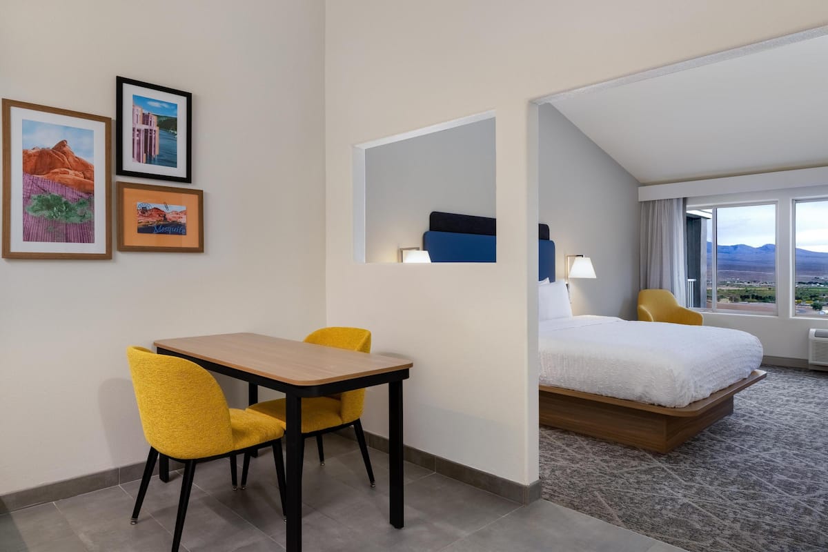 A hotel room with a bed, a desk with two chairs, a window with a view, and three framed pictures on the wall.