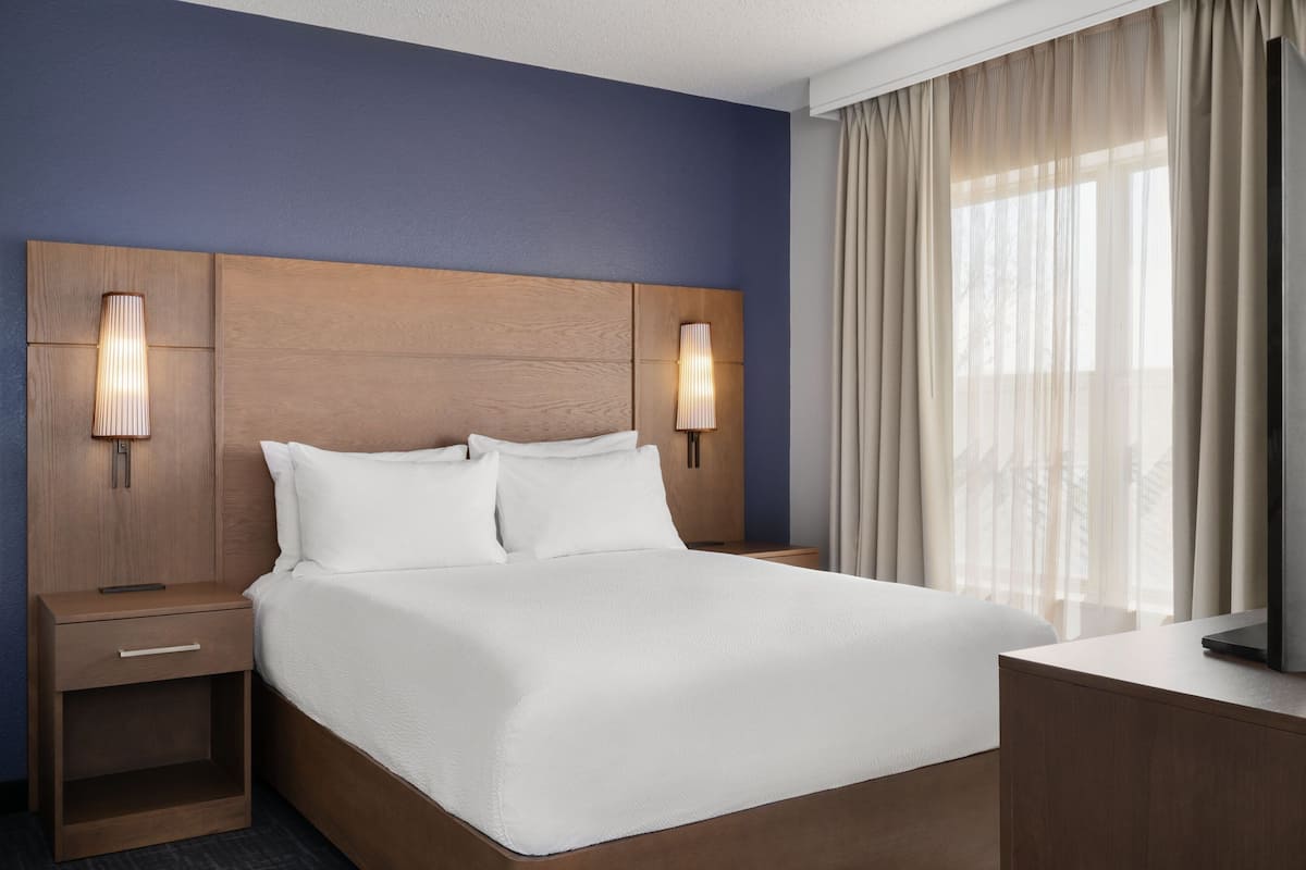 A hotel room with a large bed, wooden headboard, two bedside lamps, a TV, and a window with curtains.