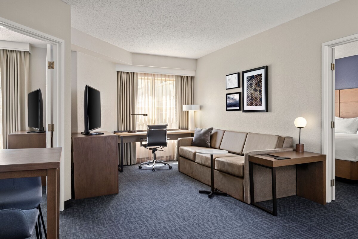 A hotel room with a desk, a sofa, a TV, a lamp, and framed pictures on the wall.