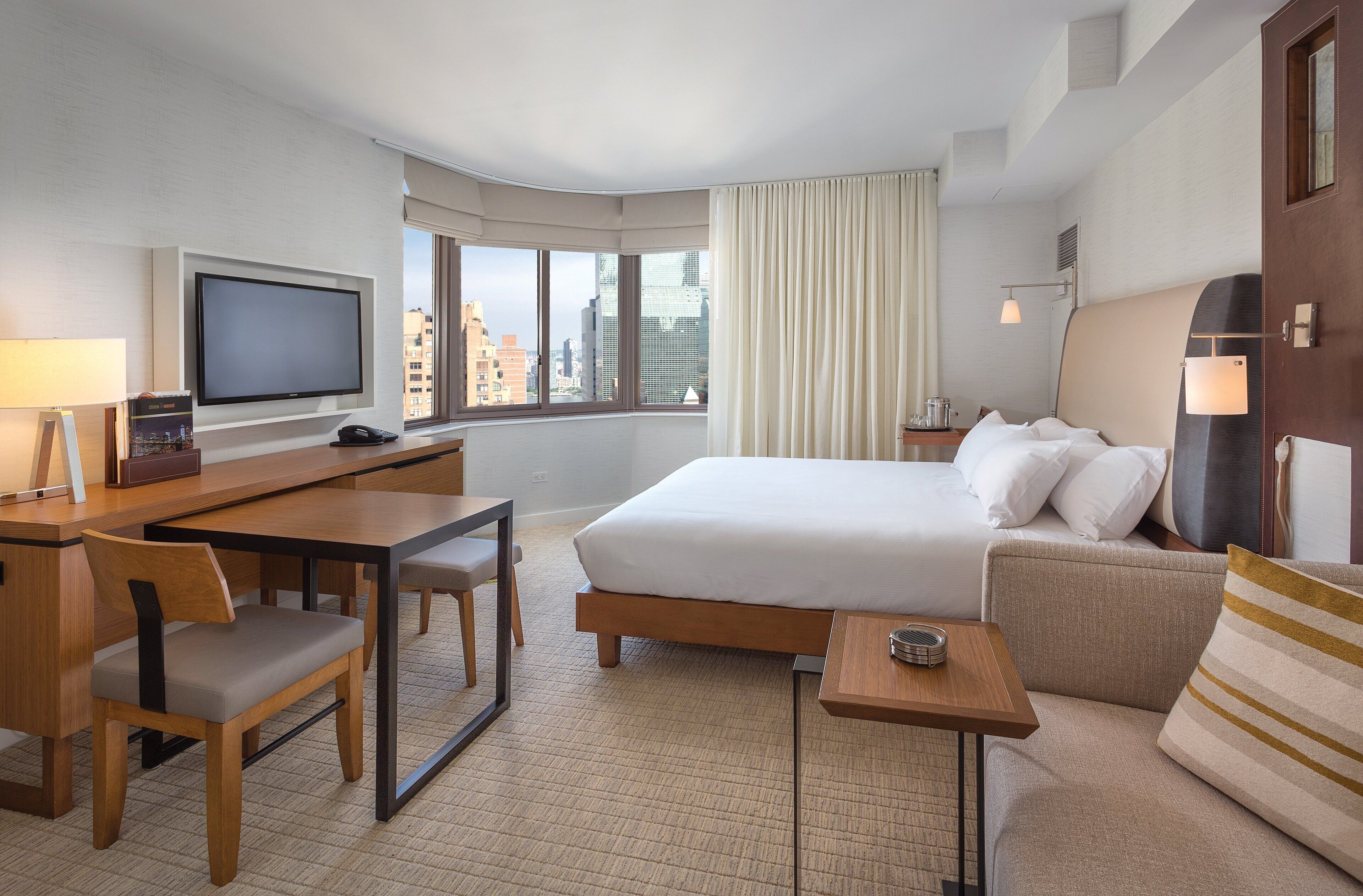 Habitación de hotel con una cama grande, un escritorio con una silla, televisión y vistas a la ciudad a través de amplios ventanales.
