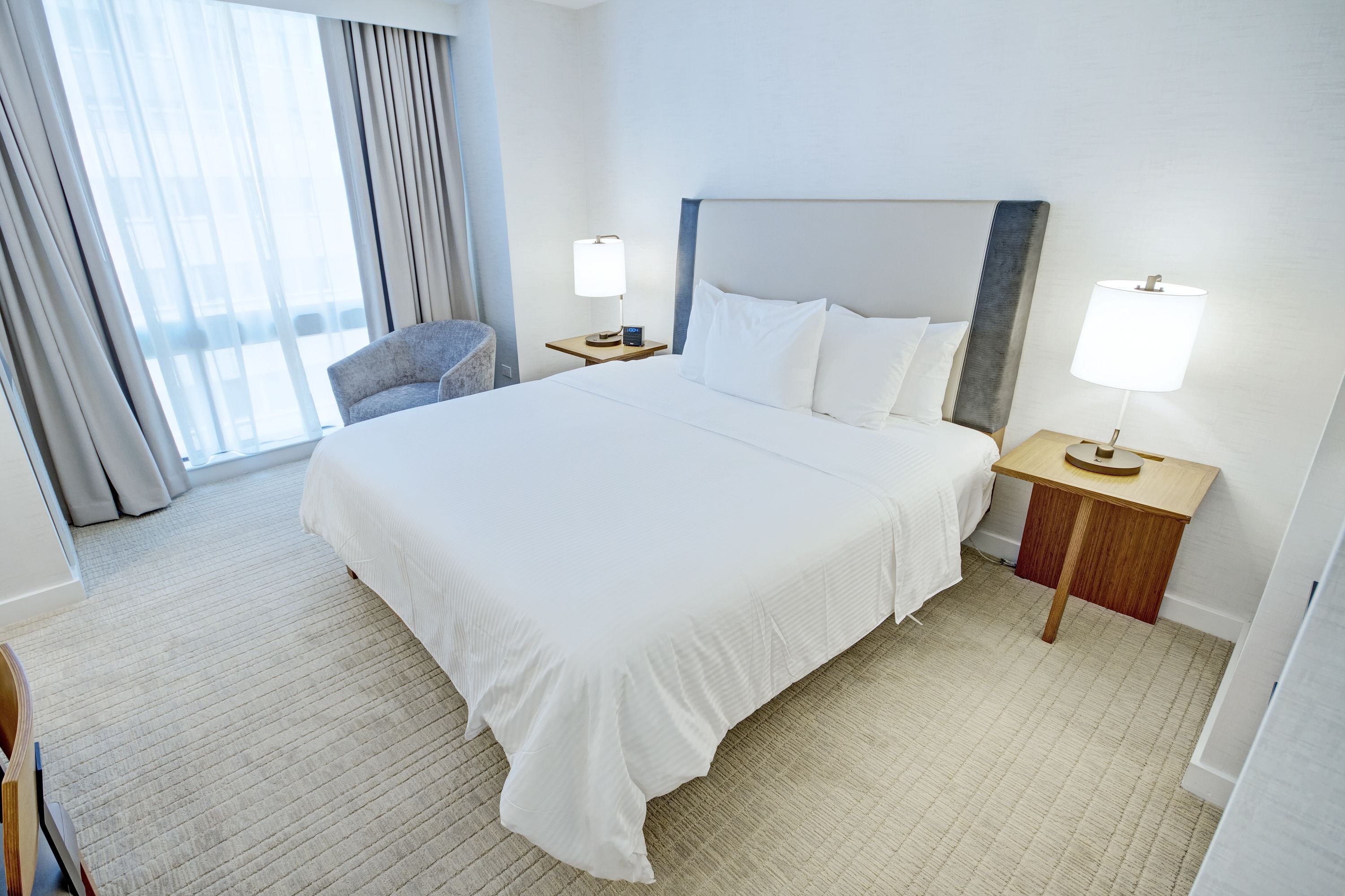 Habitación de hotel con una cama grande, dos mesitas de noche con lámparas, una silla y vistas al exterior a través de una ventana con cortinas.
