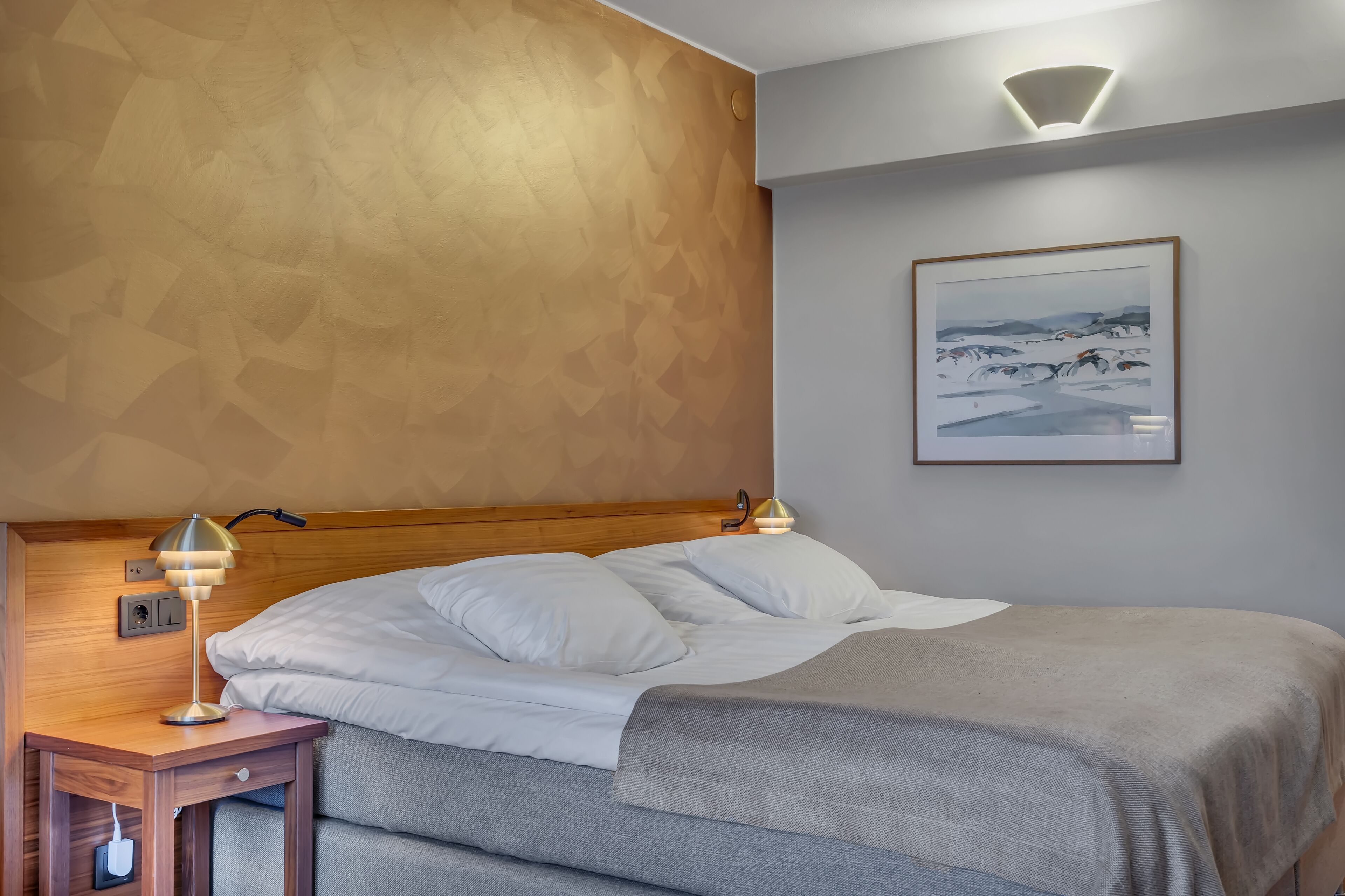 Una cama bien hecha con sábanas blancas, un plaid gris y un cabecero de madera. Hay una mesita de noche con una lámpara y un enchufe. Un cuadro enmarcado cuelga en la pared sobre la cama.
