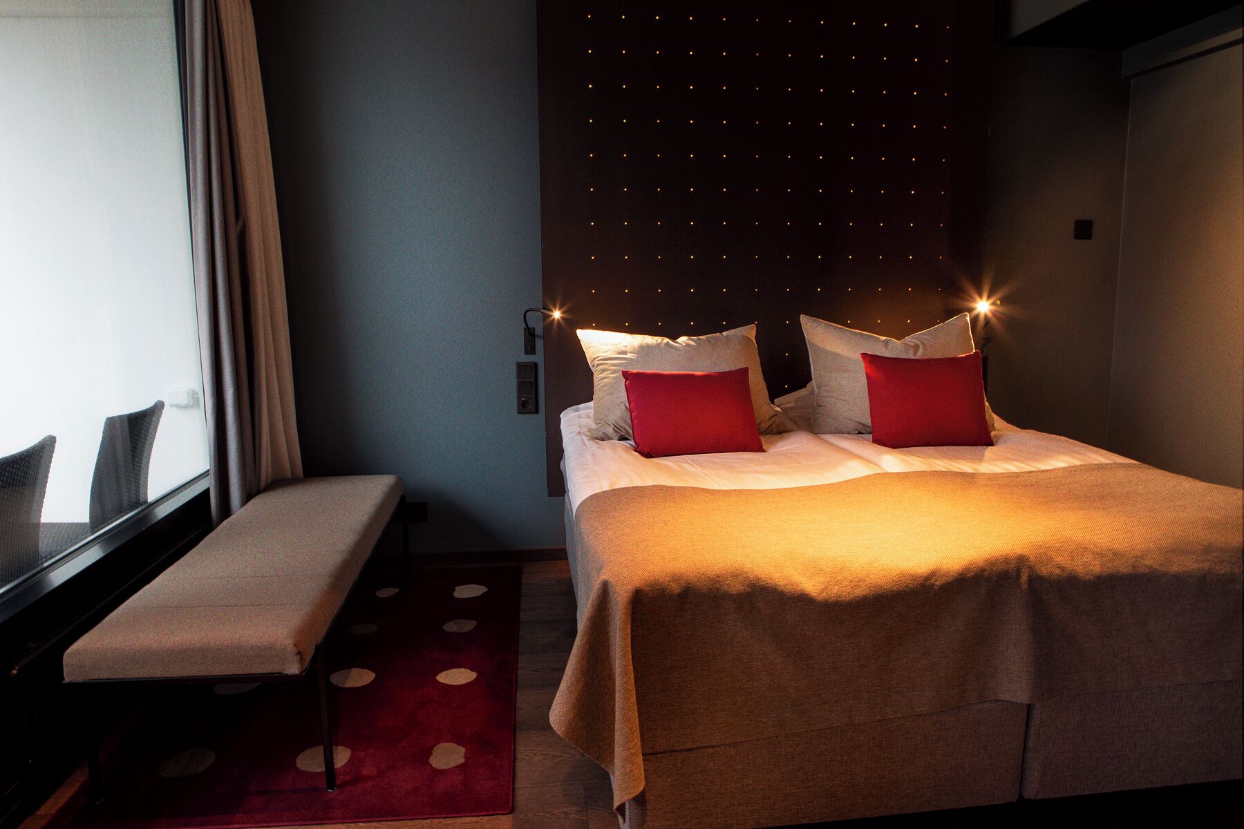 A hotel room with a bed, a bench, a wall-mounted light fixture, and a window.