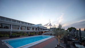 A swimming pool with a sunken deck, surrounded by lounge chairs and umbrellas, with a building featuring multiple balconies and a covered area in the background. - Hotell Arkipelag (Mariehamn)