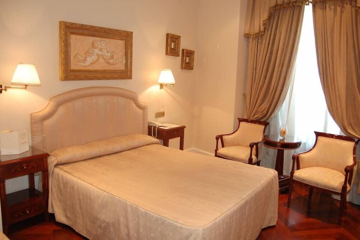 Une chambre d’hôtel avec un grand lit, deux tables de chevet, deux chaises et une lampe fixée au mur.