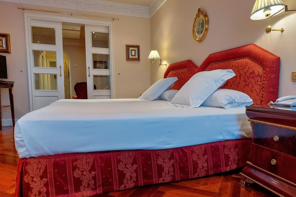 Une chambre à coucher avec un grand lit, une tête de lit rouge en tissu, une table de chevet en bois et une lampe fixée au mur.